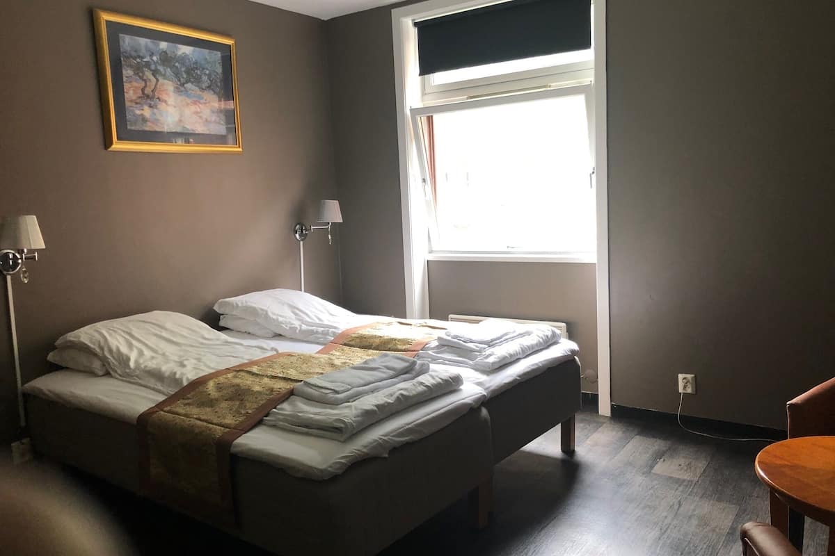 A hotel room with two beds, a painting on the wall, a window with blinds, a lamp, and a chair.