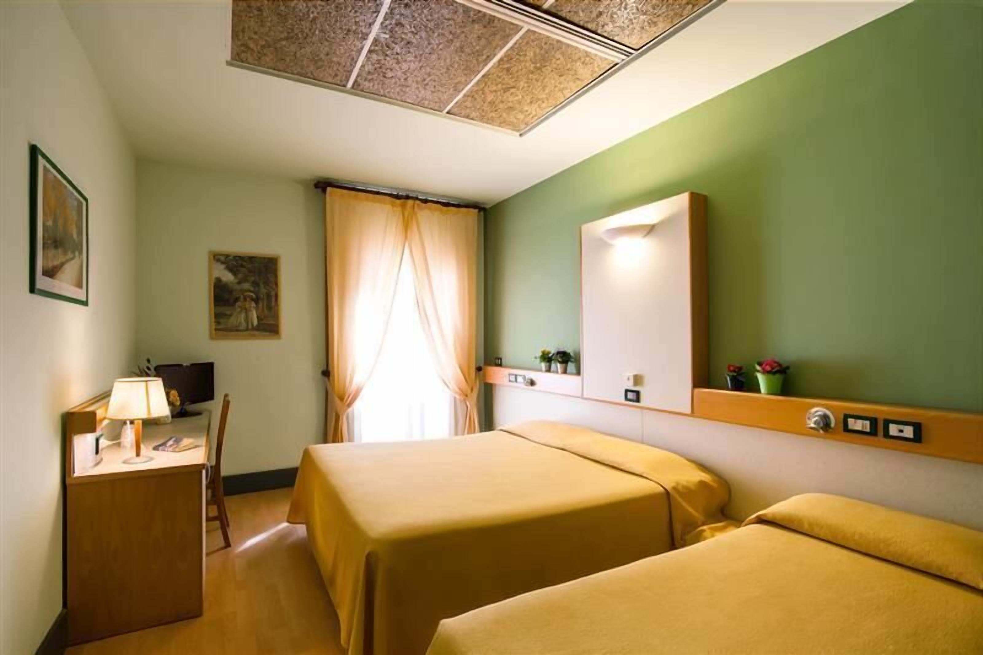 A hotel room with two beds, a desk with a lamp, a television, and a window with curtains.