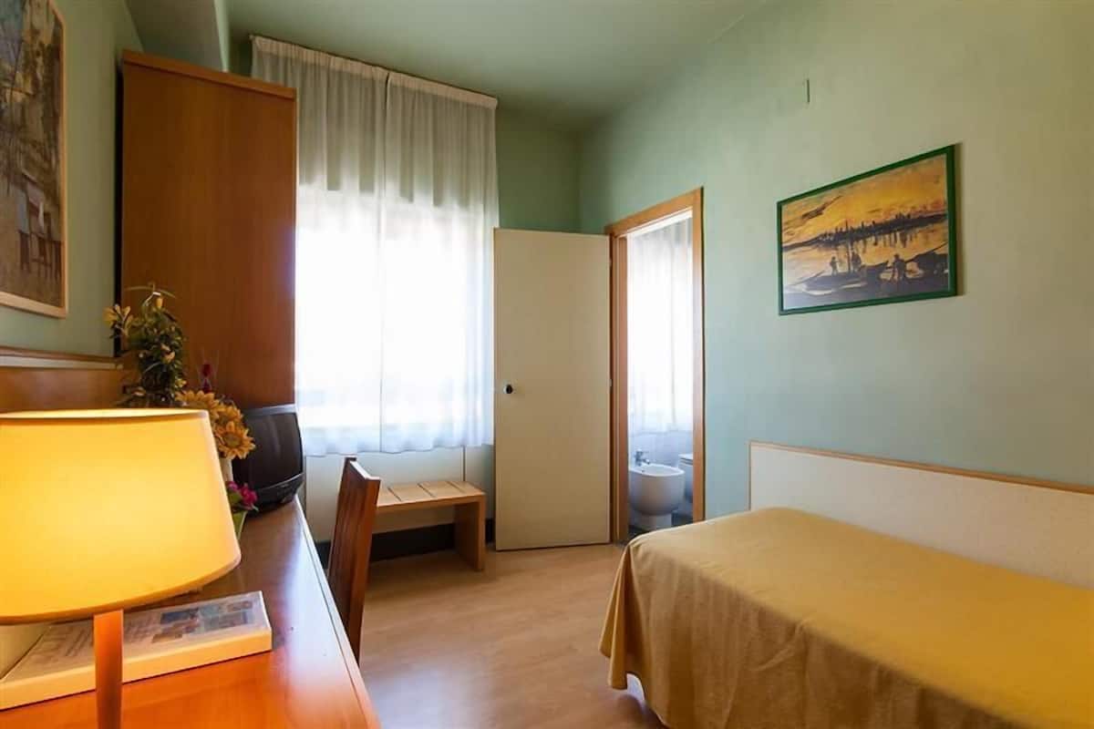 A room with a bed, a desk with a lamp, a chair, a painting on the wall, and a door leading to a bathroom.
