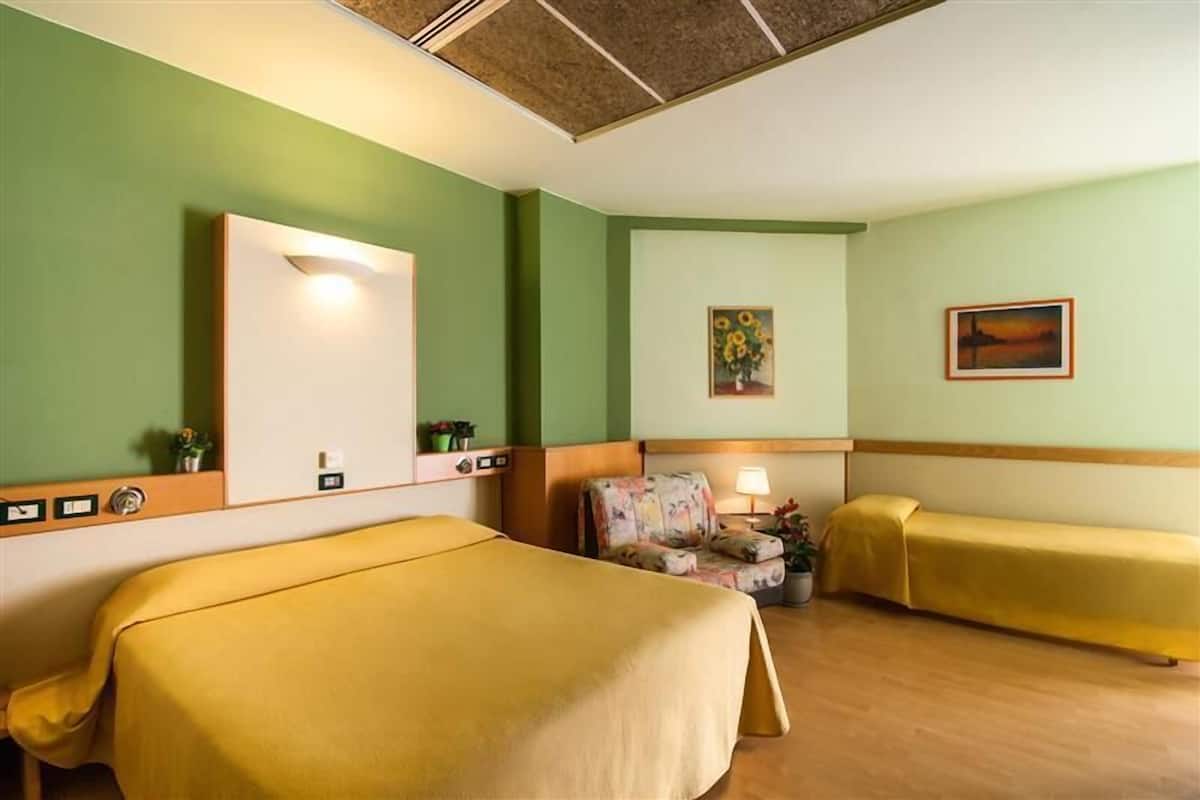 A hotel room with two beds, a chair, a small table with a lamp, and a painting on the wall.