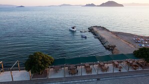 A coastal area with a boat docked near a rocky shoreline, a sandy beach with umbrellas, and a row of tables and chairs.