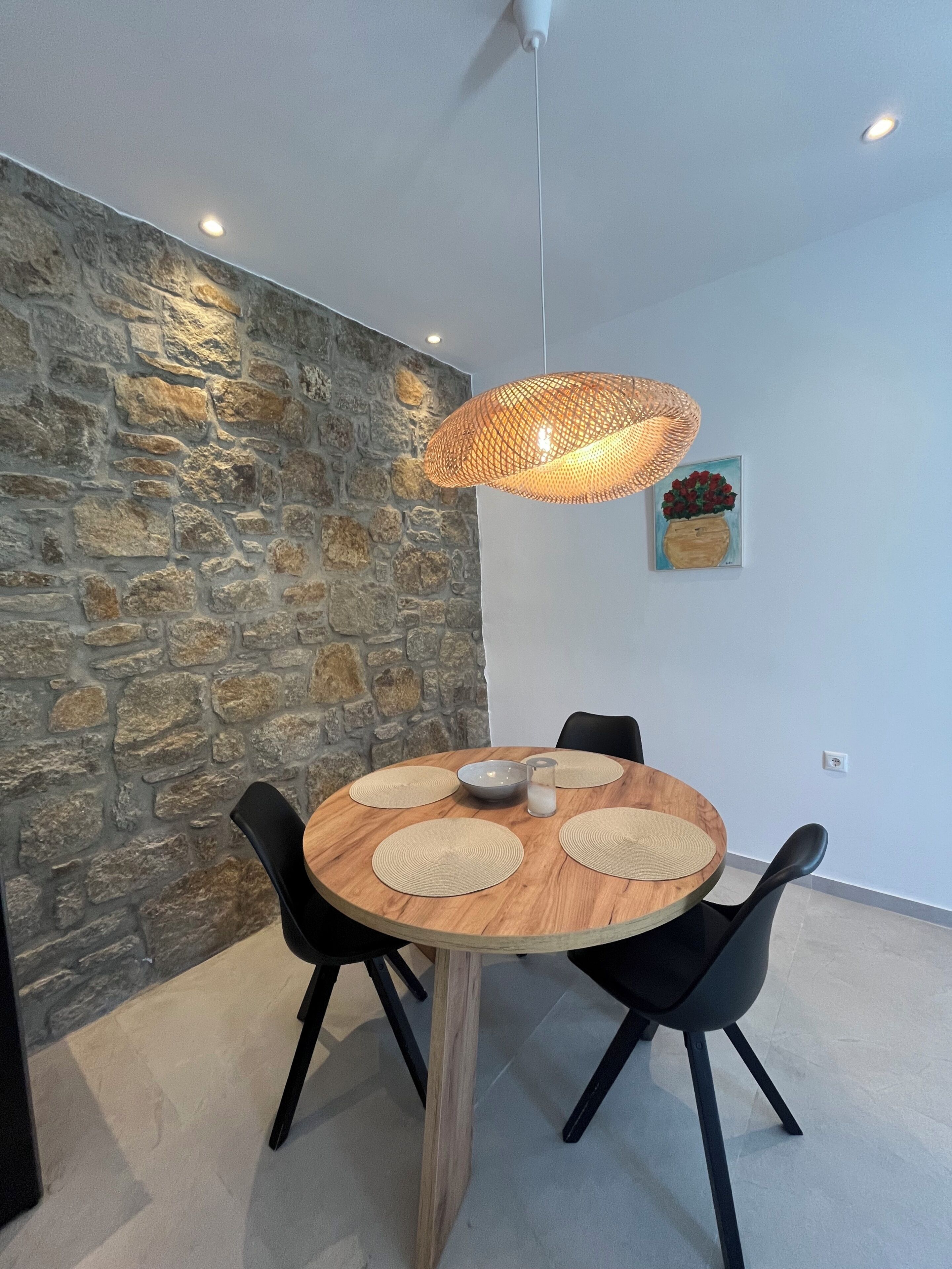 Ein Essbereich mit einem runden Holztisch, schwarzen Stühlen, einer gewobenen Pendelleuchte, einer Steinwand und einem gerahmten Gemälde.