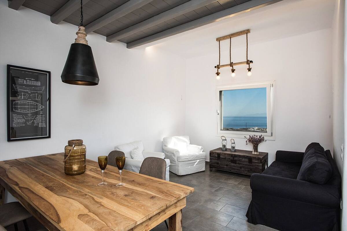A modern living room with a wooden dining table, white sofas, and a large window offering a view of the sea.