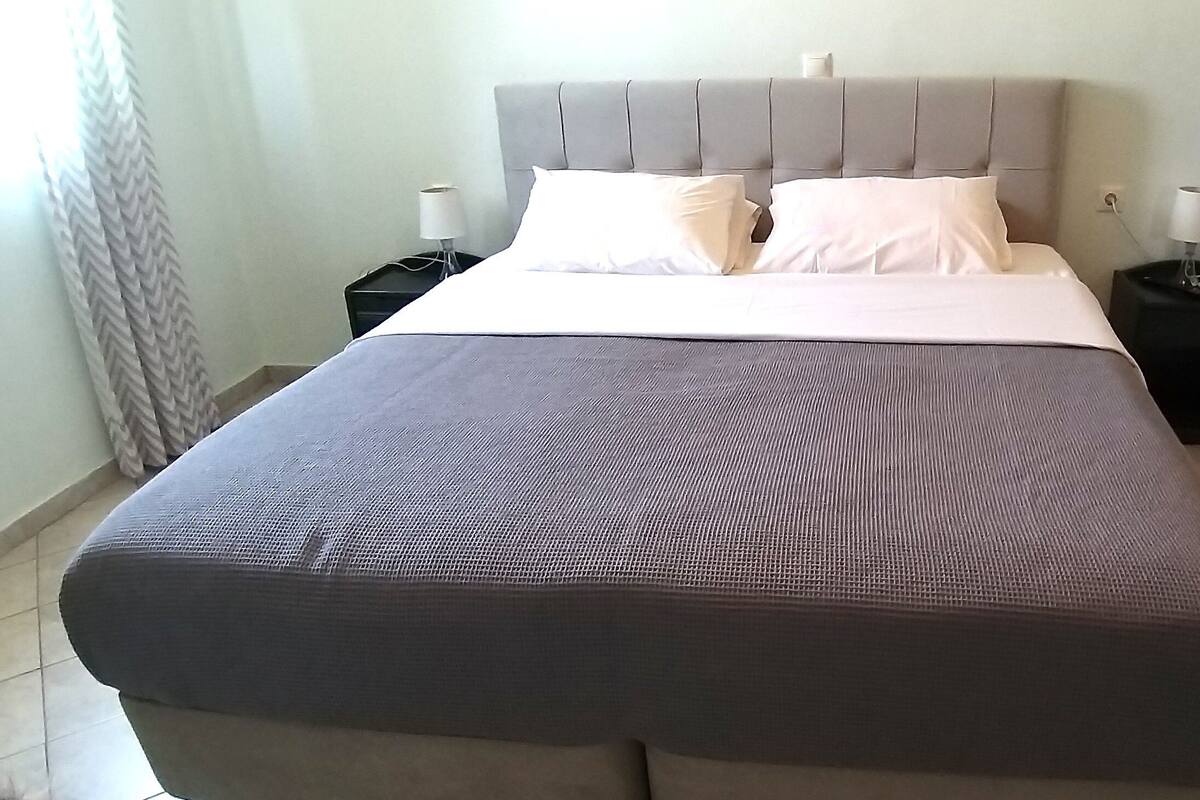 A neatly made bed with a grey headboard, two bedside tables with lamps, and a tiled floor.