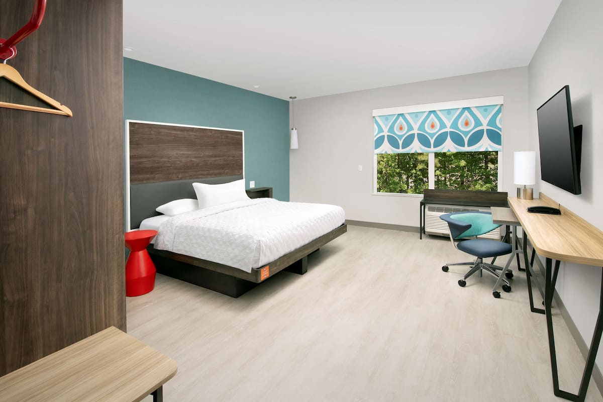 Une chambre d’hôtel moderne équipée d’un lit, d’un bureau, d’une chaise, d’une télévision et d’une fenêtre donnant sur la verdure.