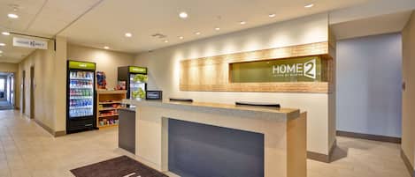 A hotel reception area with a reception desk, a 'Home 2' sign, a 'cold' beverage machine, and a 'really cold' beverage machine.