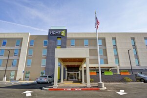 Entrada de un hotel con un letrero que dice "HOME 2 Suites by Hilton", un acceso cubierto y un asta con la bandera estadounidense.
