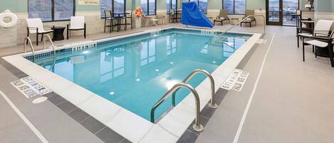 An indoor swimming pool with clear safety signage, a lifeguard chair, and a view of the outside through large windows.