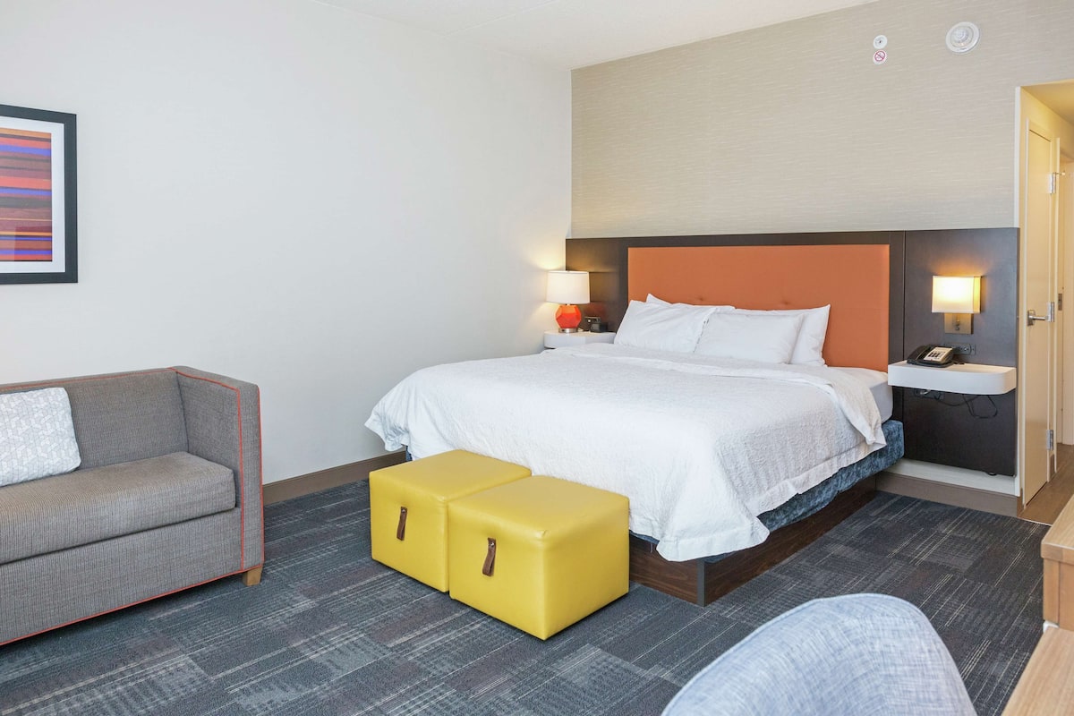 A hotel room with a bed, a sofa, two yellow ottomans, a nightstand with a lamp, and a wall-mounted phone.
