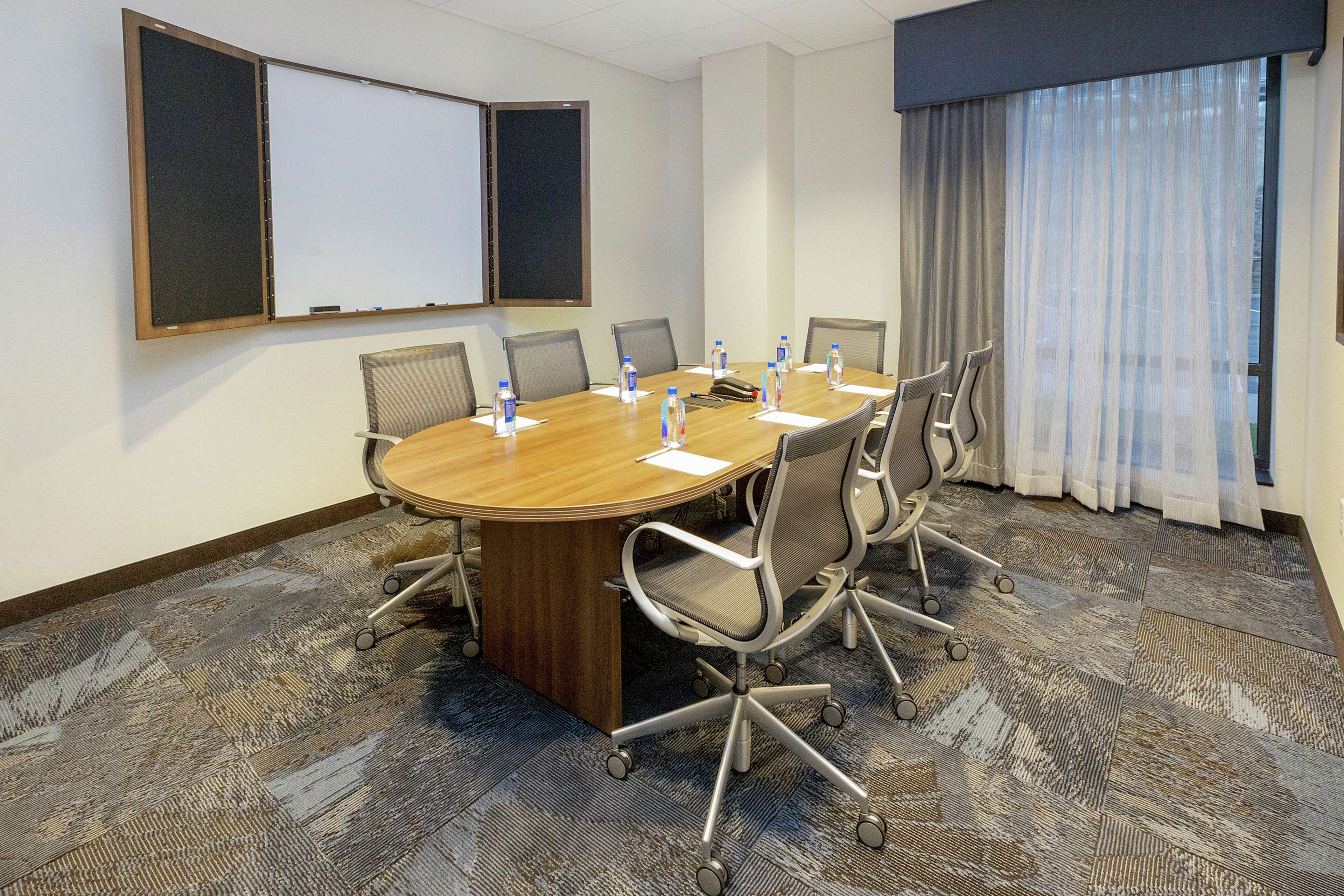A conference room with a round table, six chairs, a whiteboard, and a window with curtains.