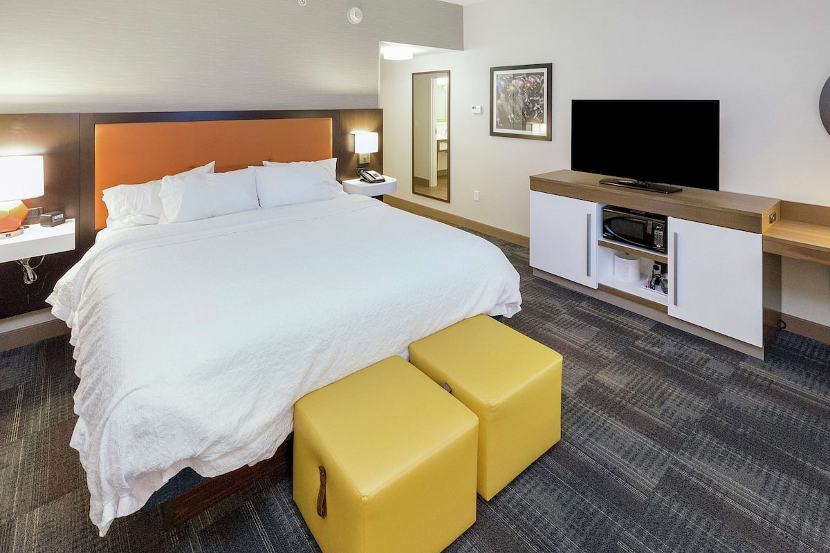 A hotel room with a large bed, a bedside table with a lamp, a TV, a mirror, and a minibar.