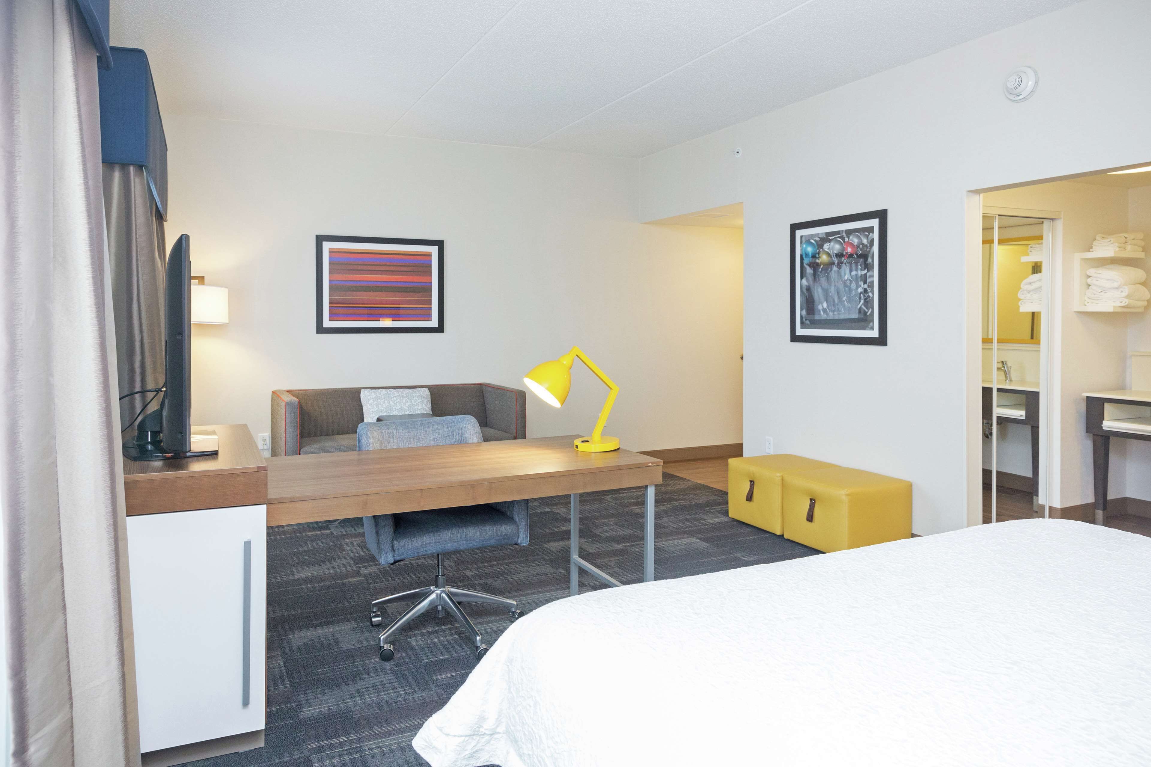 A hotel room with a bed, a desk with a chair, a desk lamp, a small yellow ottoman, a television, and artwork on the walls.
