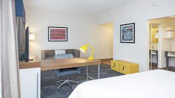 A hotel room with a bed, a desk with a chair, a desk lamp, a small yellow ottoman, a television, and artwork on the walls.