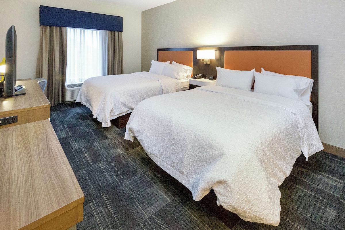 A hotel room with two beds, a desk, a TV, and a window with curtains.