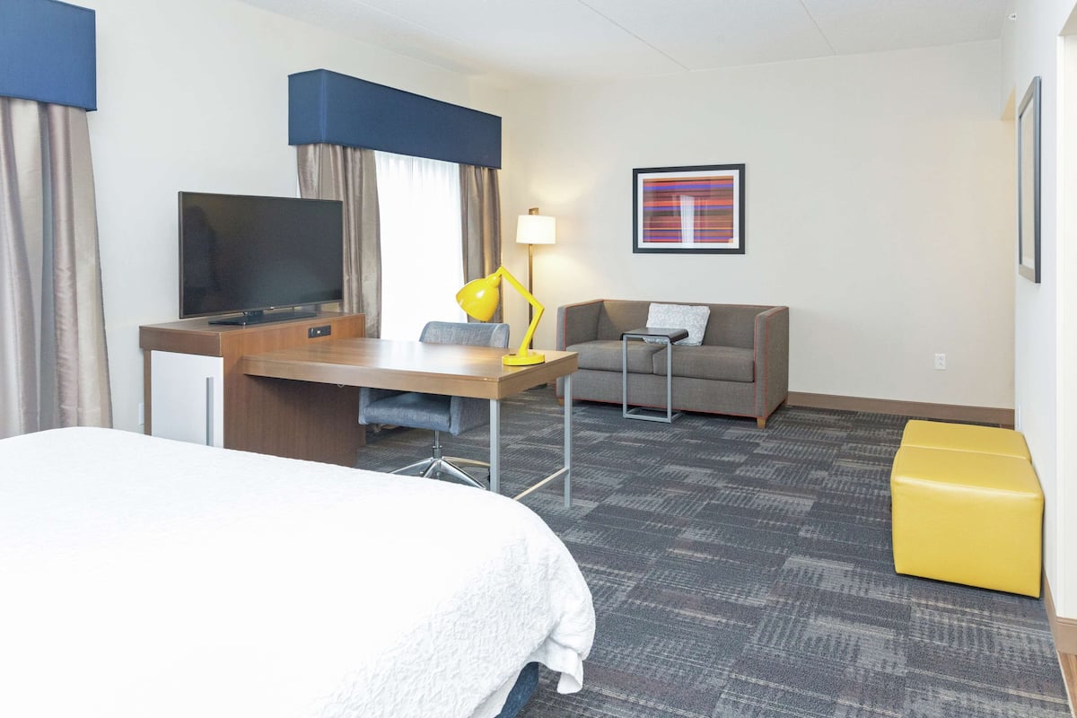 A hotel room with a bed, a desk with a TV, a lamp, a sofa, and a small table.