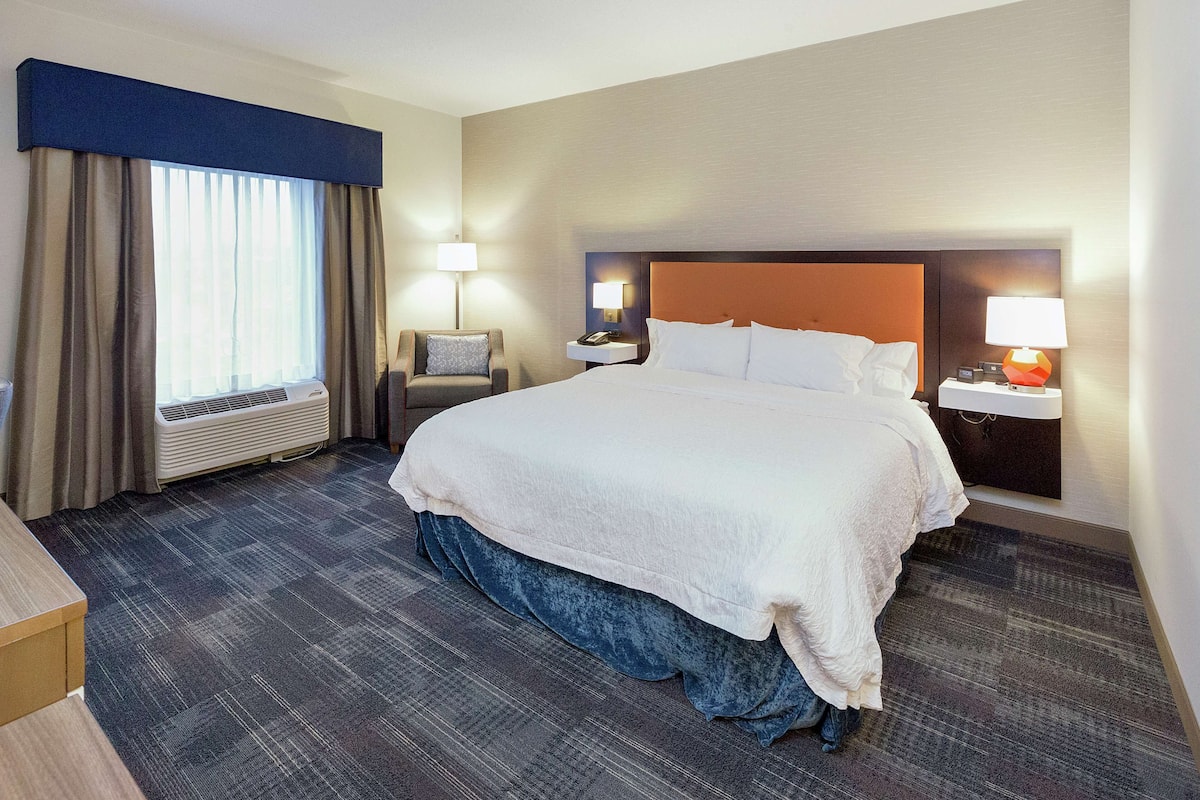 A hotel room with a large bed, a desk, a chair, a nightstand with a lamp, and a window with curtains.