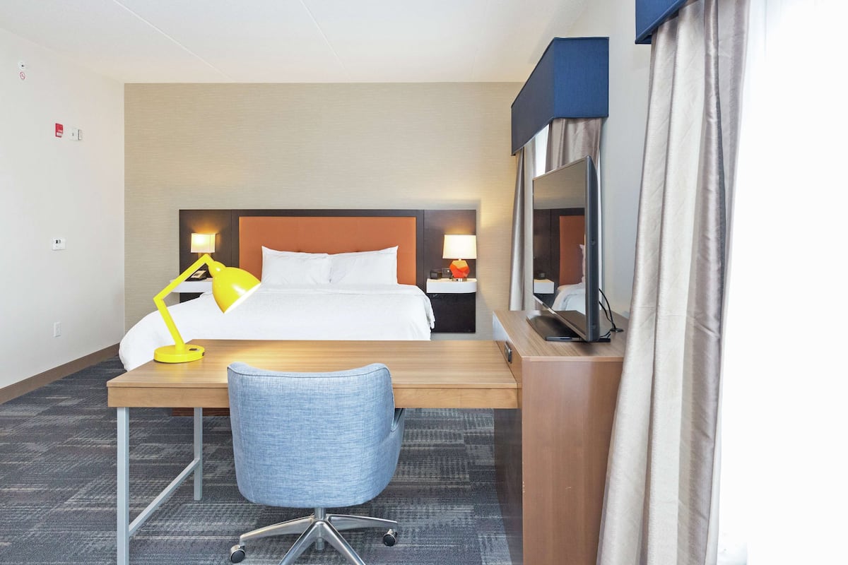 A hotel room with a bed, desk, chair, two bedside lamps, a mirror, and a window with curtains.