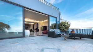 A modern outdoor patio area with a seating arrangement, a large window, and a view of the sky. - Villa Azure (Vari-Voula-Vouliagmeni)