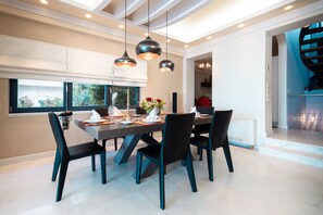 Un espace repas moderne avec une table rectangulaire dressée pour six personnes, des chaises noires, un éclairage suspendu et une vue sur une cuisine et un escalier.