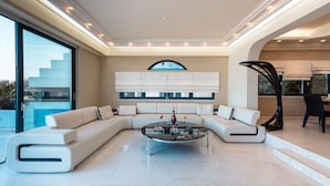 A modern living room with a sectional sofa, a glass coffee table, and a large window with a view. - Villa Azure (Vari-Voula-Vouliagmeni)