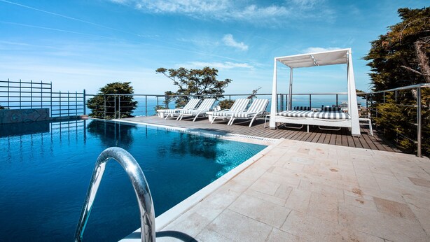 A poolside area with lounge chairs and a canopy, overlooking a body of water. - Villa Azure (Vari-Voula-Vouliagmeni)