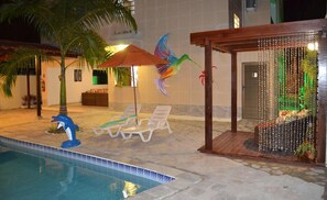 A swimming pool area with lounge chairs, a palm tree, and a decorative wall with a colorful bird. - Pousada Beija-Flores (Ipojuca)
