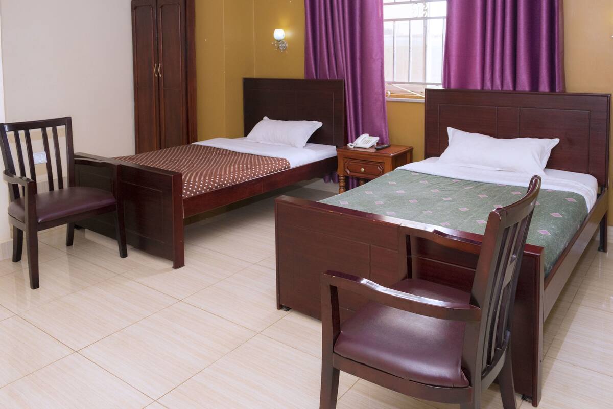 A hotel room with two wooden beds, a wooden chair, a small bedside table with a phone, and a wall-mounted light fixture.