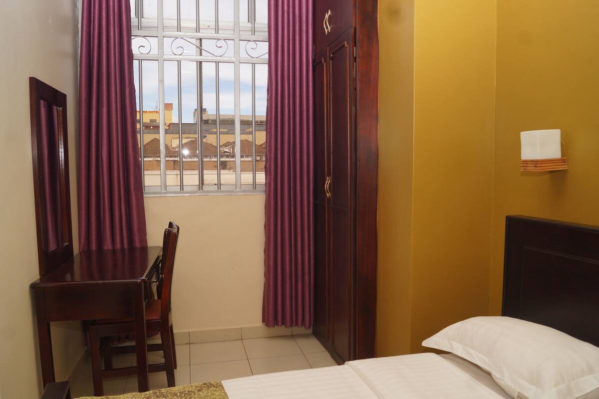 A room with a bed, a desk, and a chair. There is a window with metal bars and purple curtains.