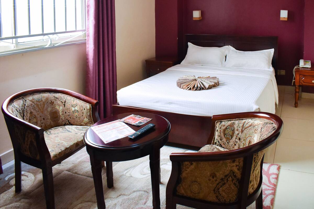 A hotel room with a bed, two armchairs, a small table with a remote control and magazine, and a nightstand with a phone.