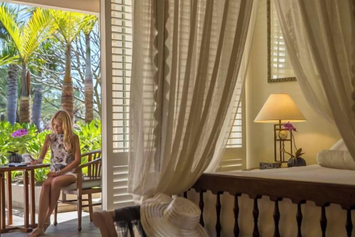 A room with a bed, a table, and a chair, with a view of greenery outside.