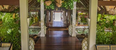 A covered walkway with wooden flooring, flanked by white pillars and wicker furniture, leading to a central area with hanging baskets and a thatched roof structure.
