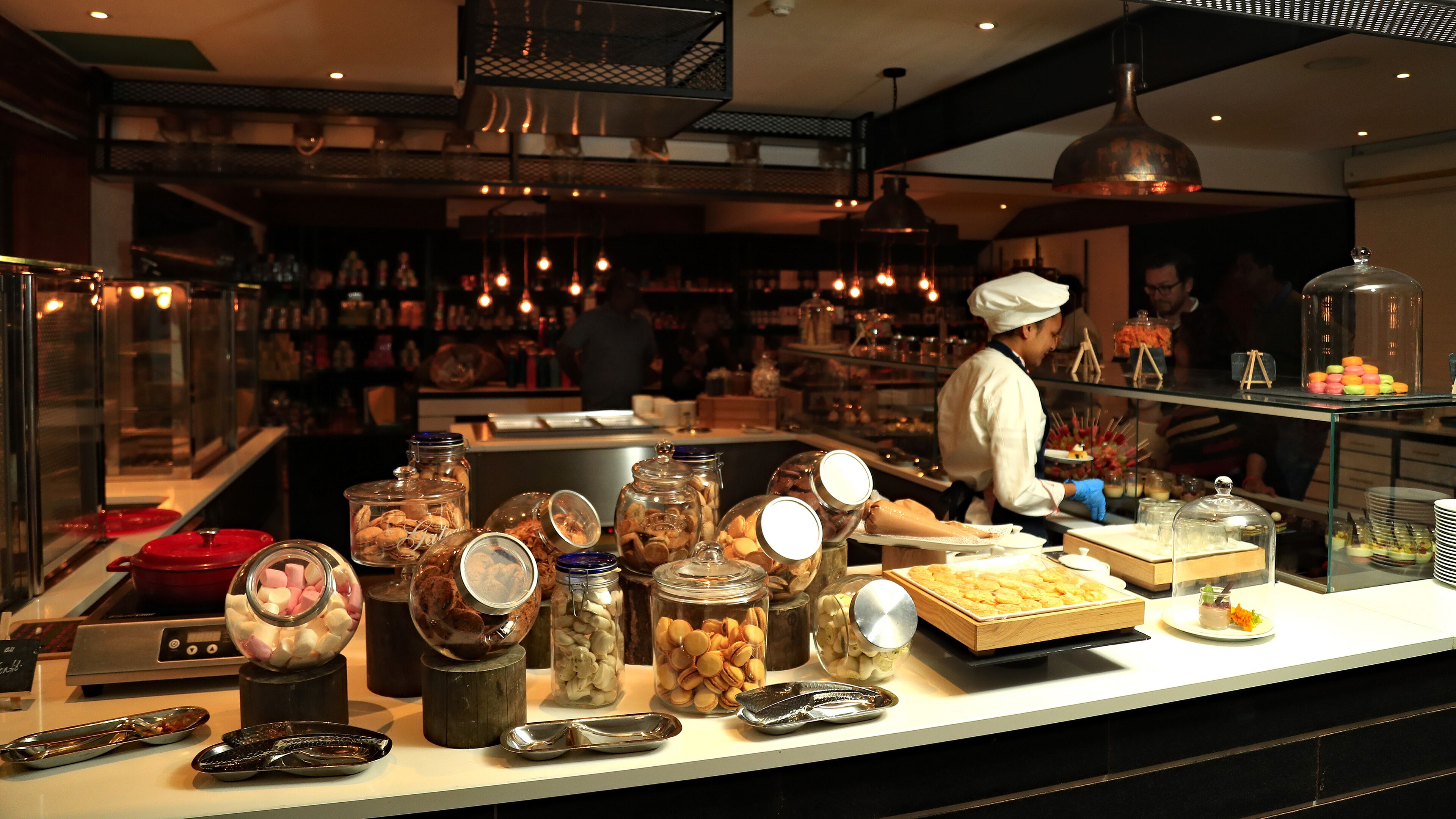 Un espace buffet où diverses préparations culinaires sont présentées dans des bocaux et des récipients, un chef qui prépare les plats, et un intérieur bien éclairé grâce à des luminaires suspendus.
