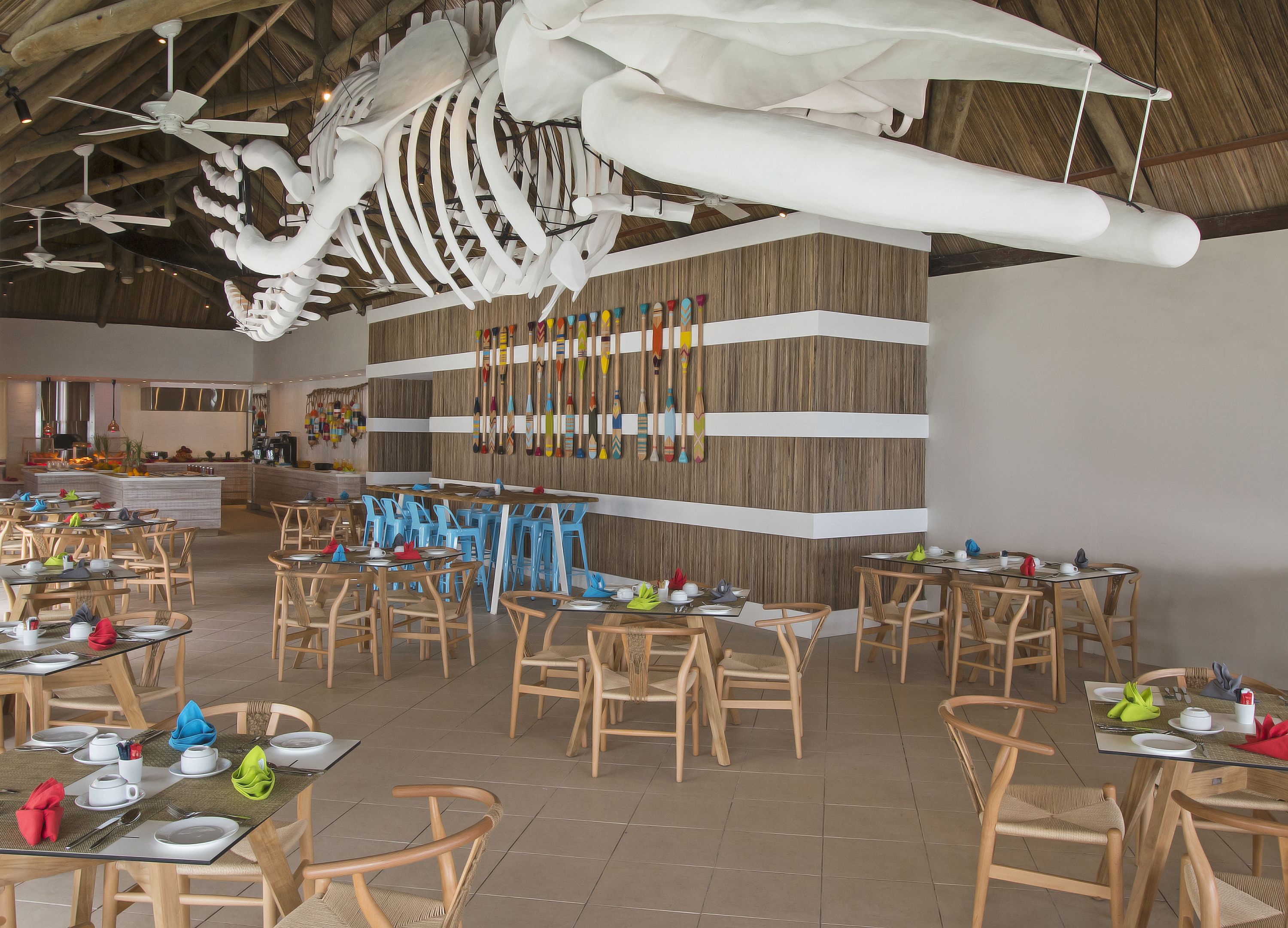 A restaurant with wooden tables and chairs, a large white sculpture on the ceiling, and a wall with colorful utensils.
