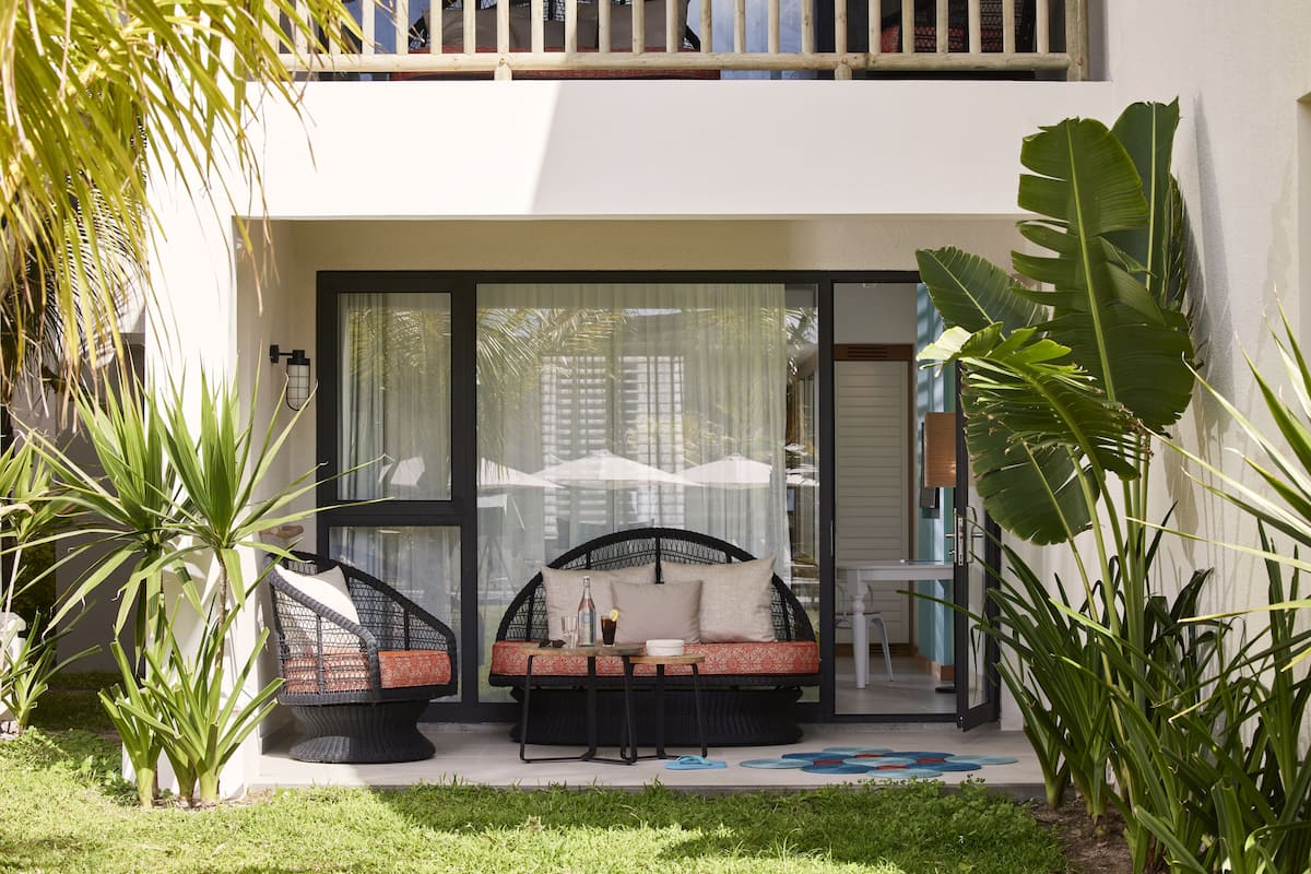 A modern outdoor seating area with a sofa and chairs, surrounded by lush greenery and a glass door leading to a balcony.