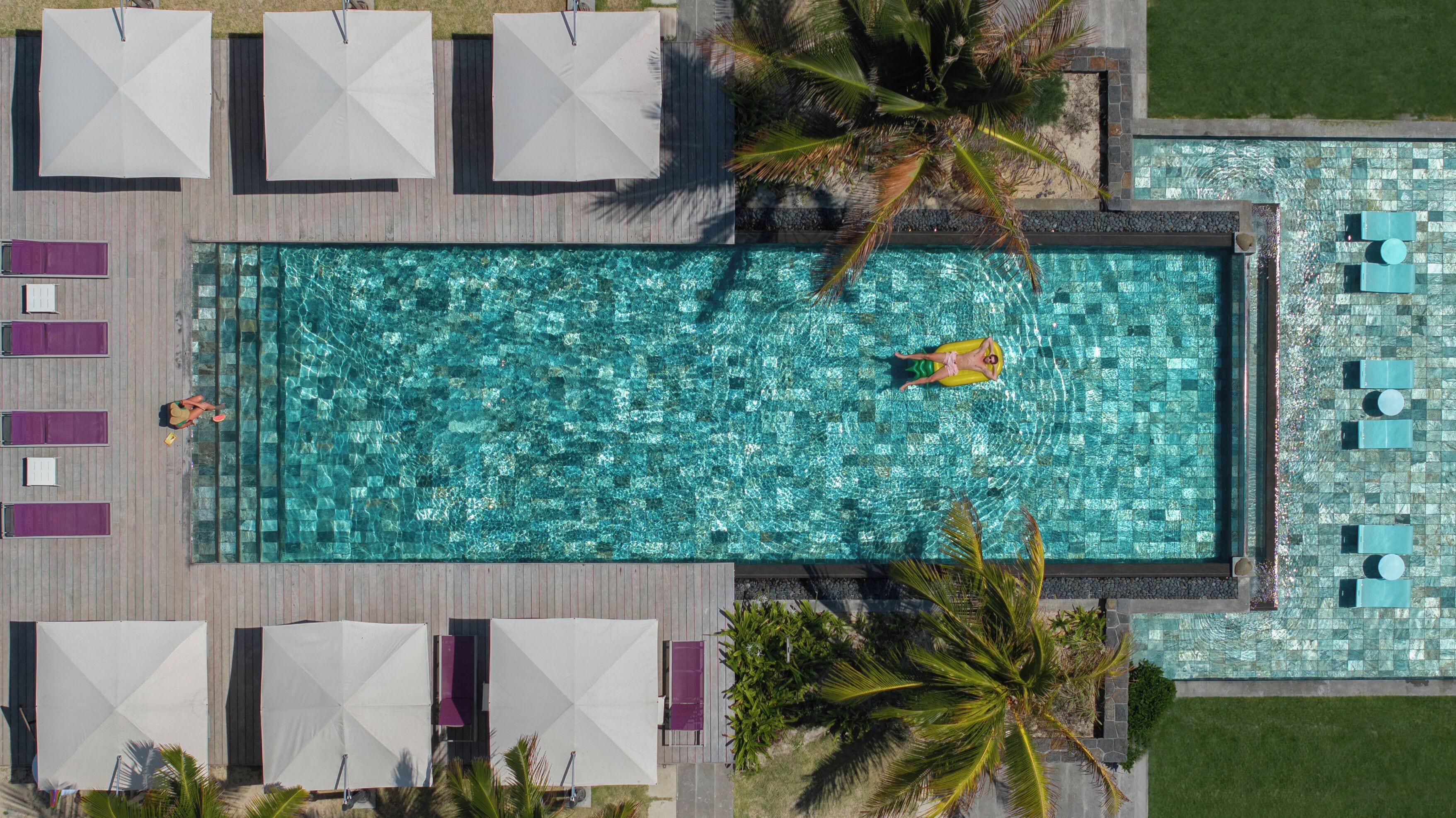 Vue aérienne d’une piscine rectangulaire entourée d’une terrasse carrelée, comprenant des chaises longues et des parasols.