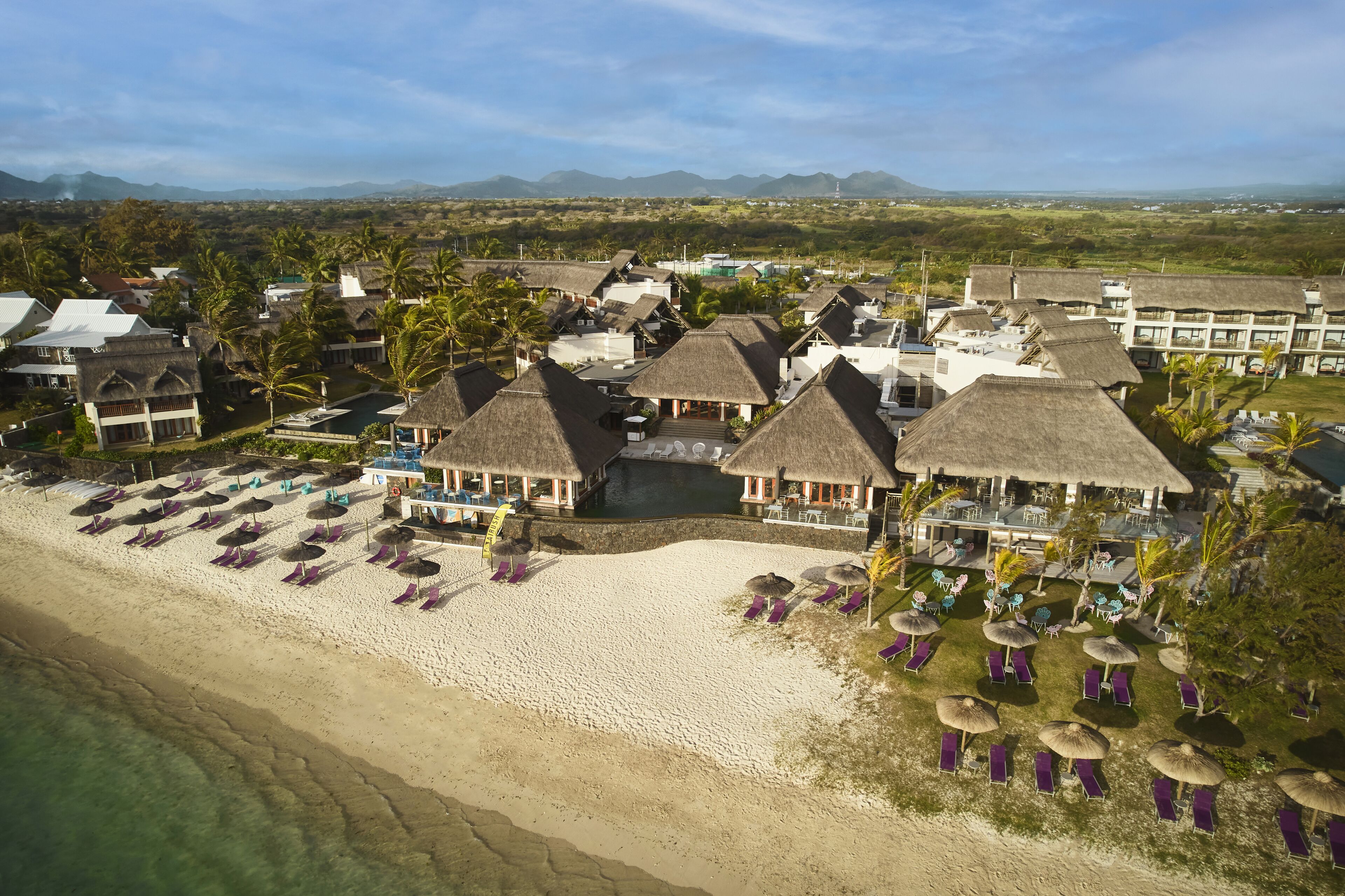 An aerial view of a resort with thatched-roof buildings, a sandy beach, and a pool area.