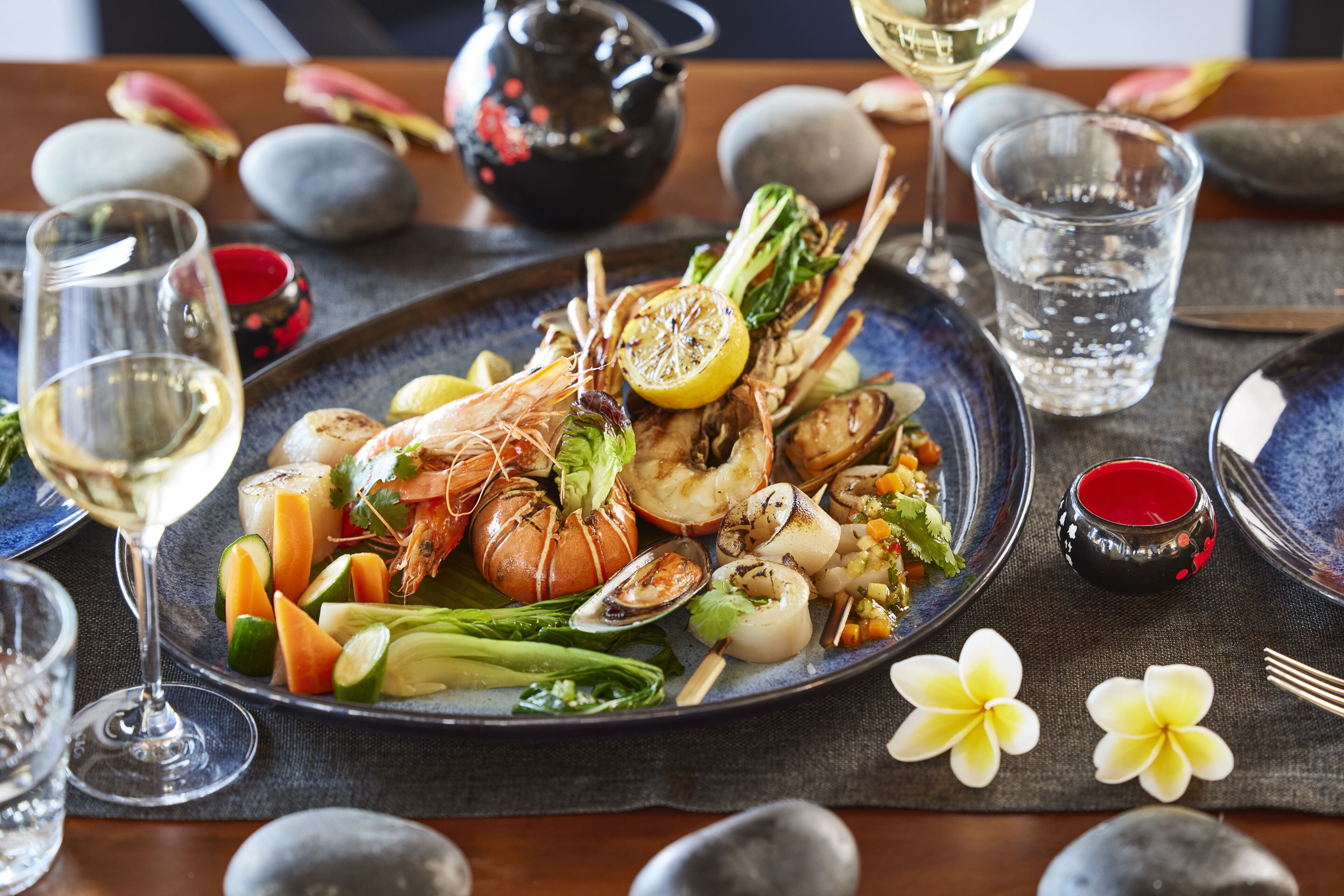 A seafood dish on a plate with various vegetables, accompanied by a glass of white wine and a small red cup.