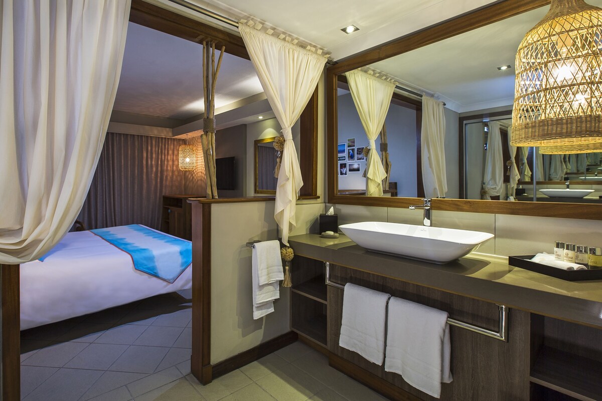 A hotel room with a bed, a large mirror, a bathroom with a sink, and a window with curtains.