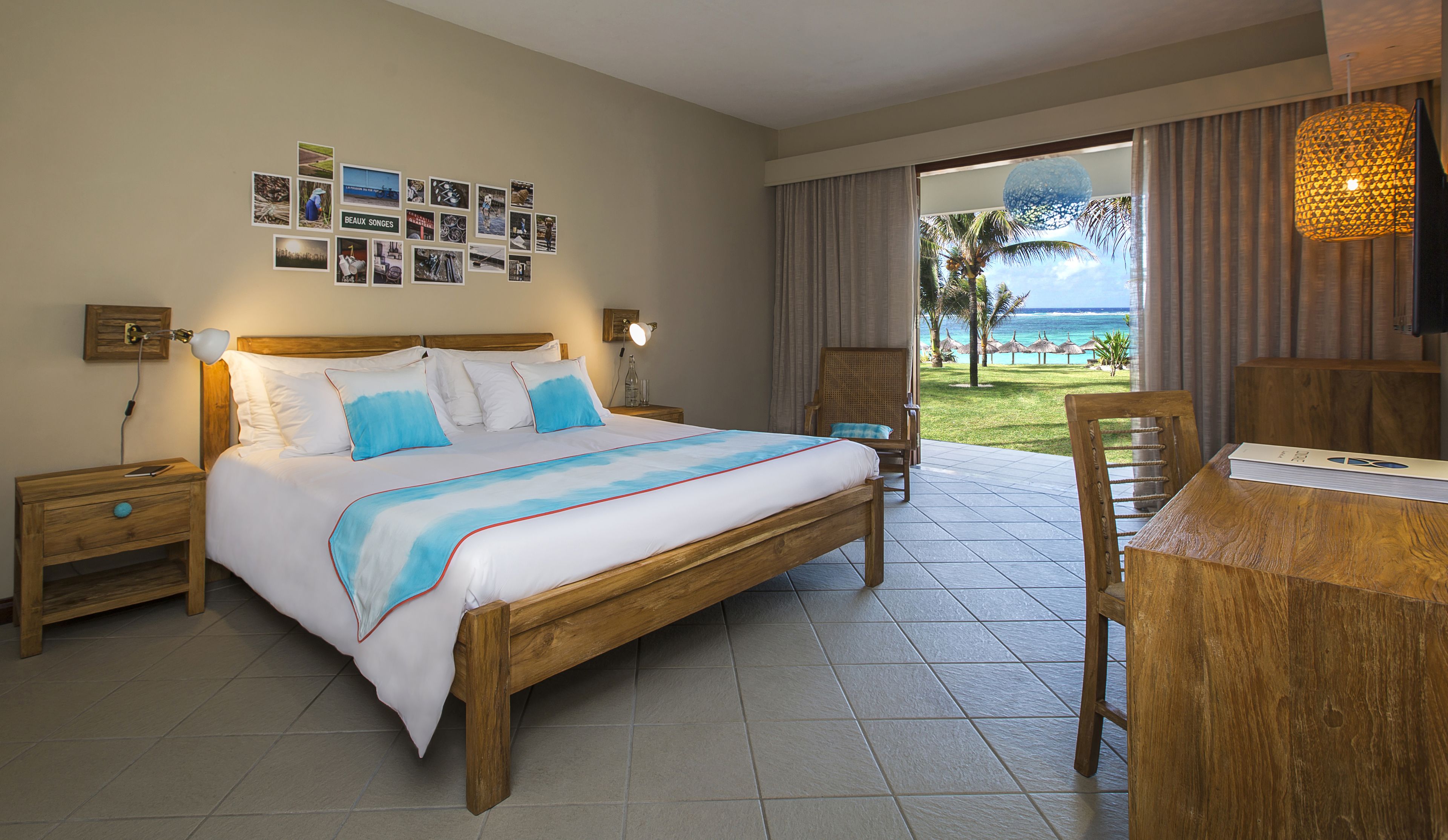 A hotel room with a bed, a desk, a chair, a nightstand, a wall with photos, and a view of the ocean.