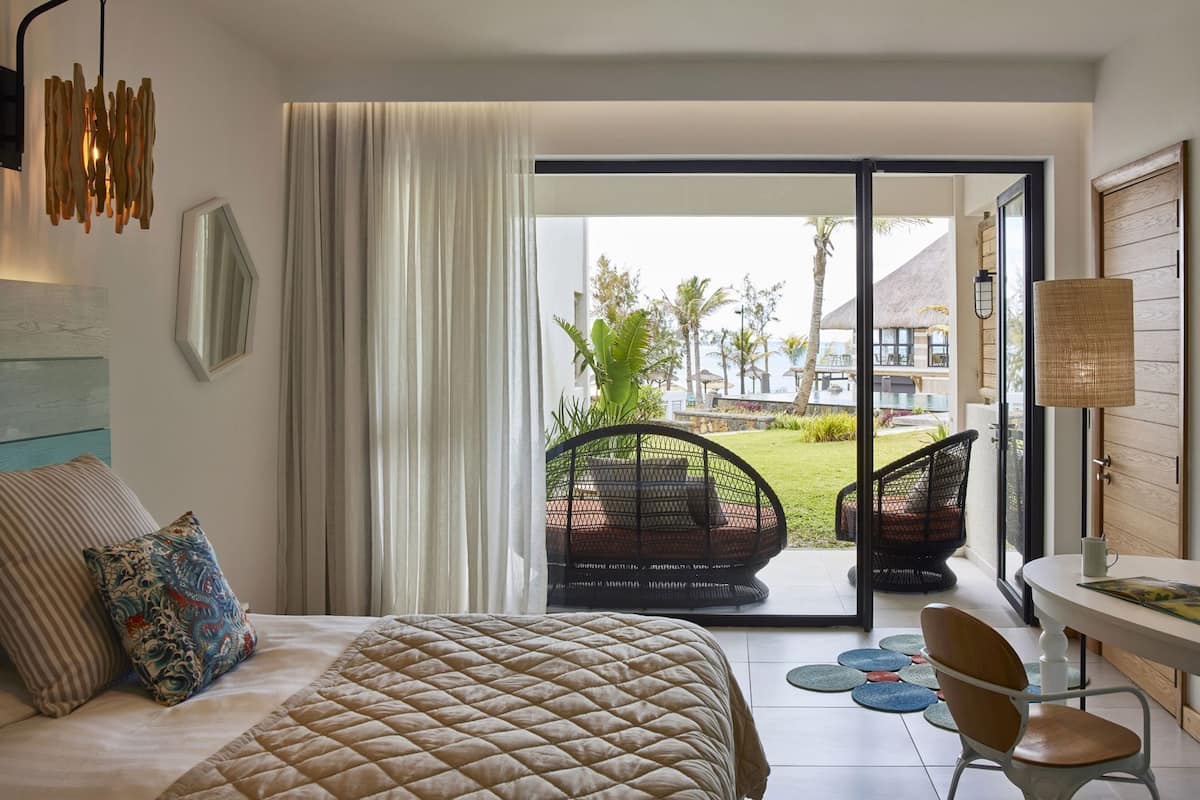 A bedroom with a bed, a chair, a table, and a view of a garden and building outside.
