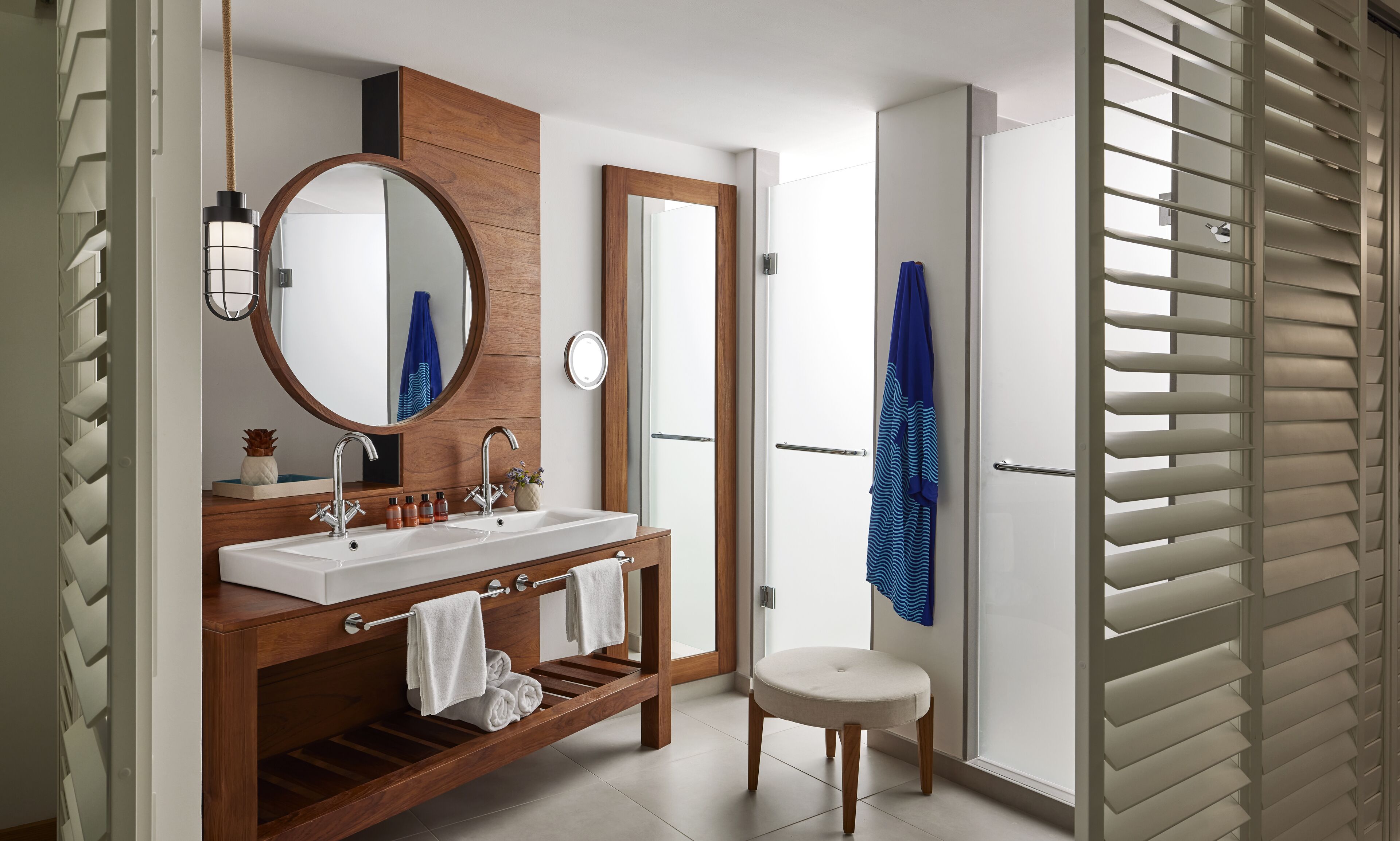 A modern bathroom with a wooden vanity, two sinks, a round mirror, and a small stool.
