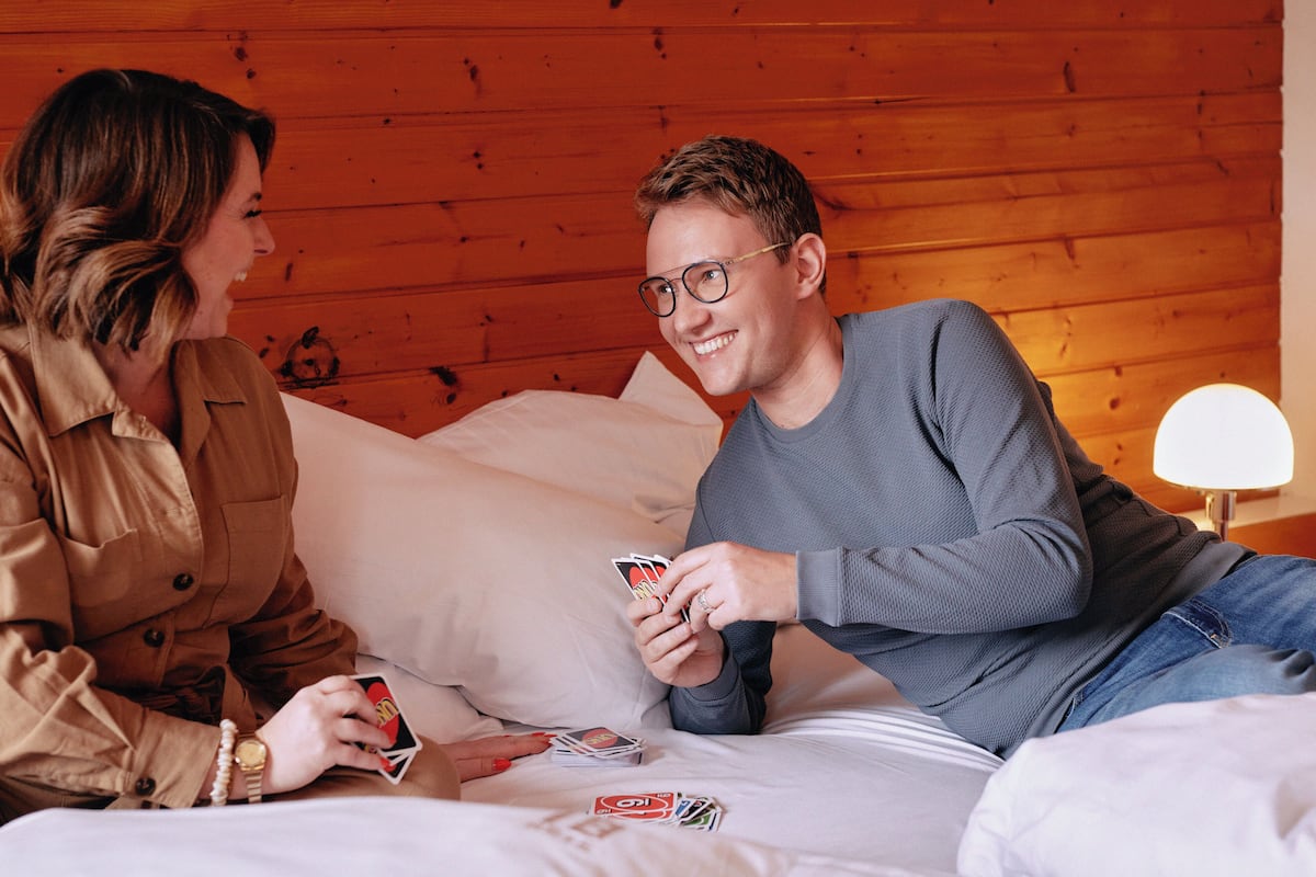 Two people sitting on a bed in a room with wooden walls, playing cards.