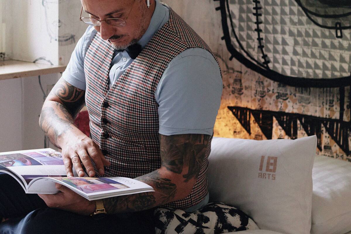 A man sitting on a bed reading a book, with a wall mural behind him.