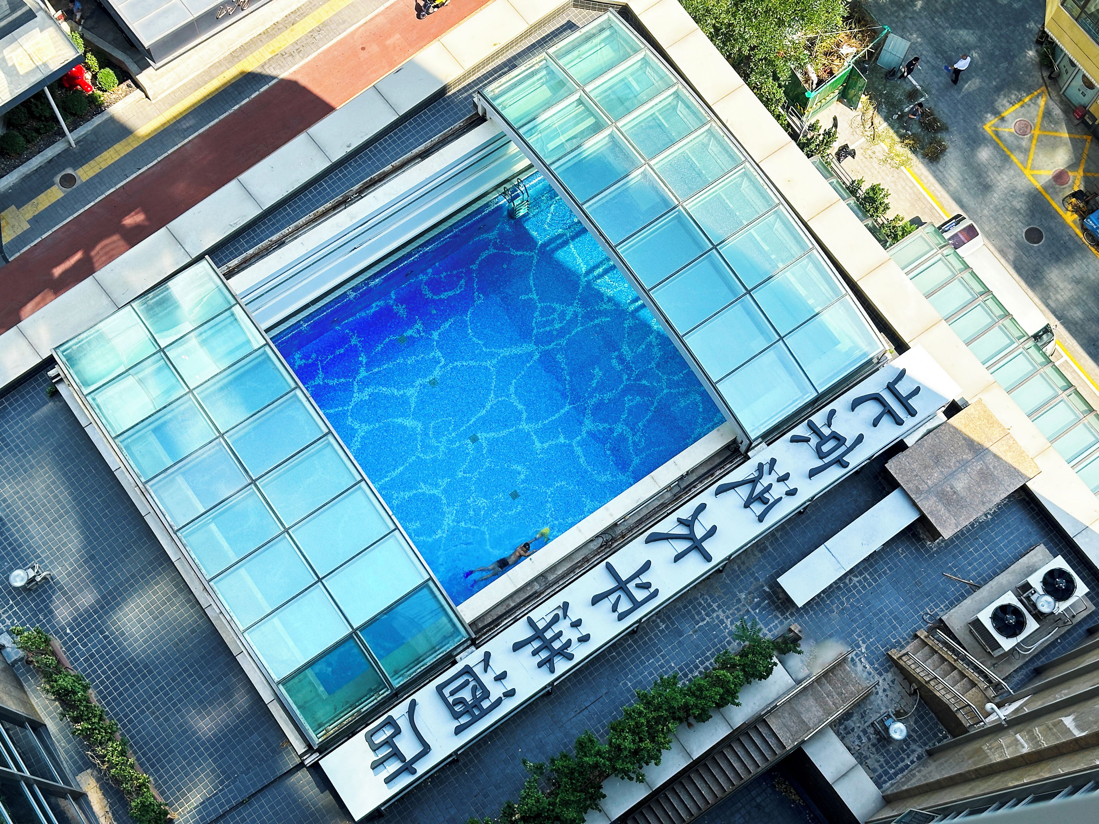 Vue aérienne d’un bâtiment doté d’une grande piscine à fond de verre, entouré d’une zone pavée et de verdure.
