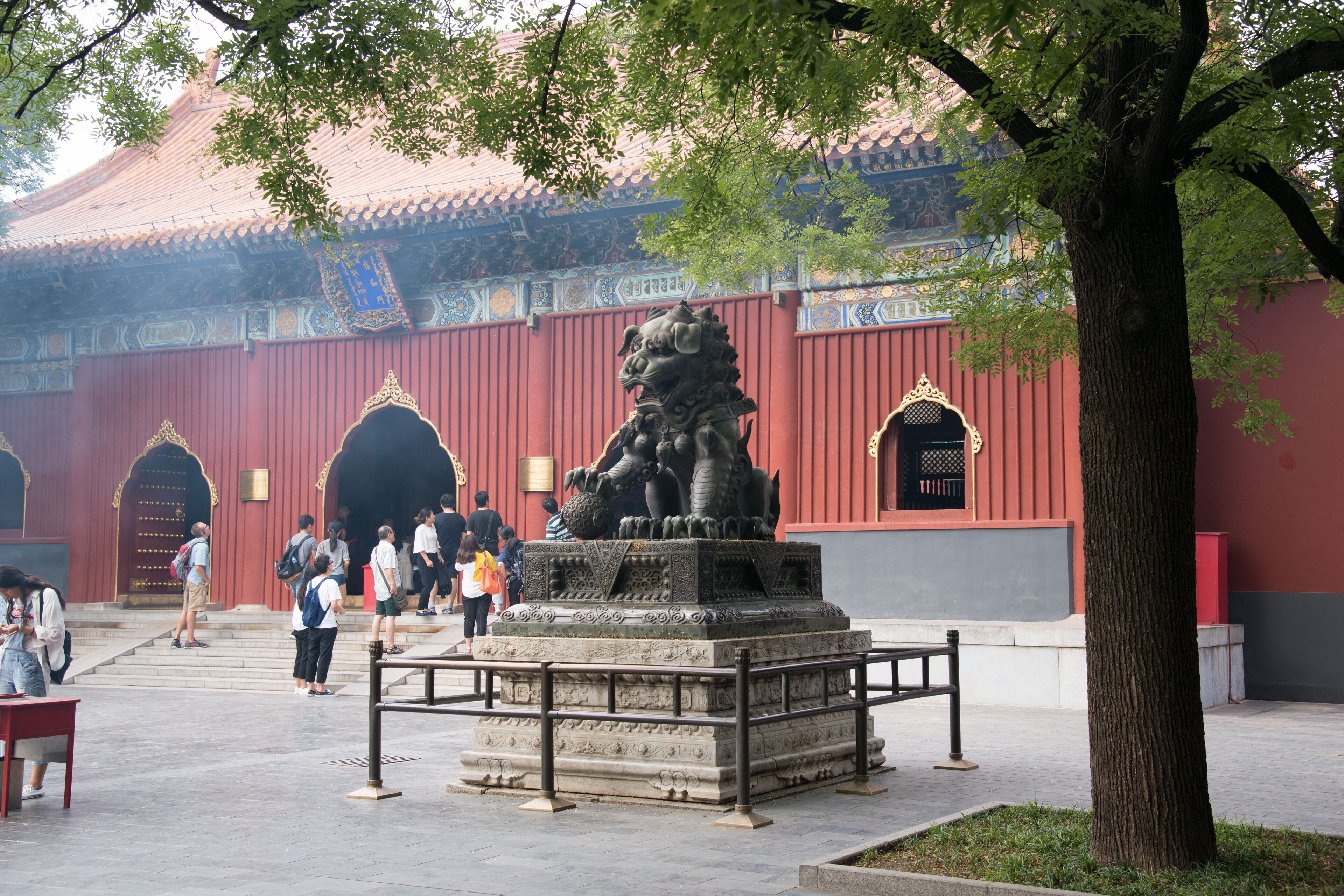A traditional building with red walls and ornate decorations, a stone lion statue, and a tree in the foreground.