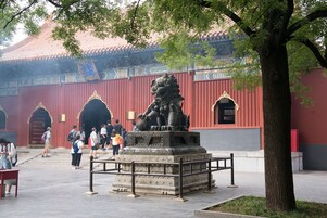 A traditional building with red walls and ornate decorations, a stone lion statue, and a tree in the foreground.