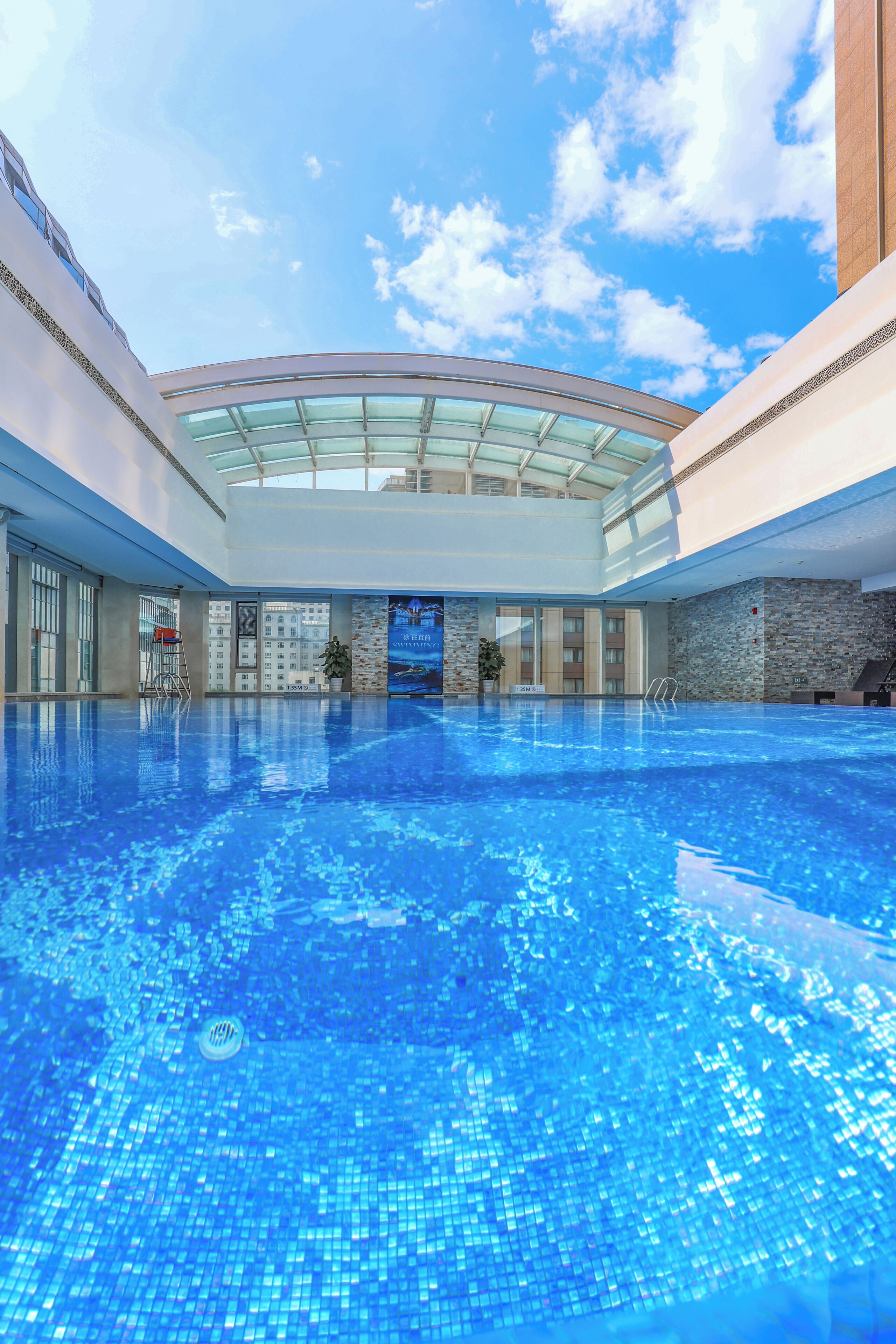 A large indoor swimming pool with a glass ceiling, surrounded by modern architecture and a clear sky.