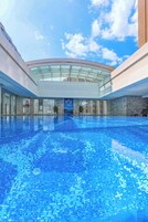 A large indoor swimming pool with a glass ceiling, surrounded by modern architecture and a clear sky.