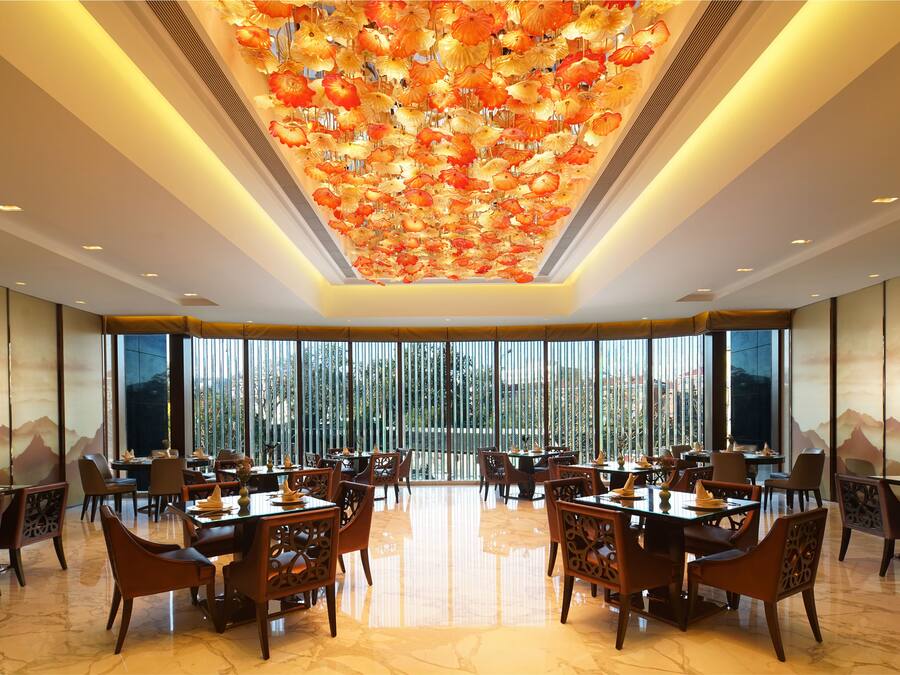 A spacious dining area with high ceilings and a decorative ceiling feature. Tables are set with chairs, and large windows provide a view of the outdoors.