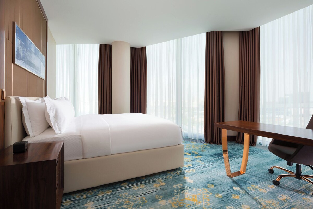 A hotel room with a bed, a desk, a chair, a large window with curtains, and a painting on the wall.